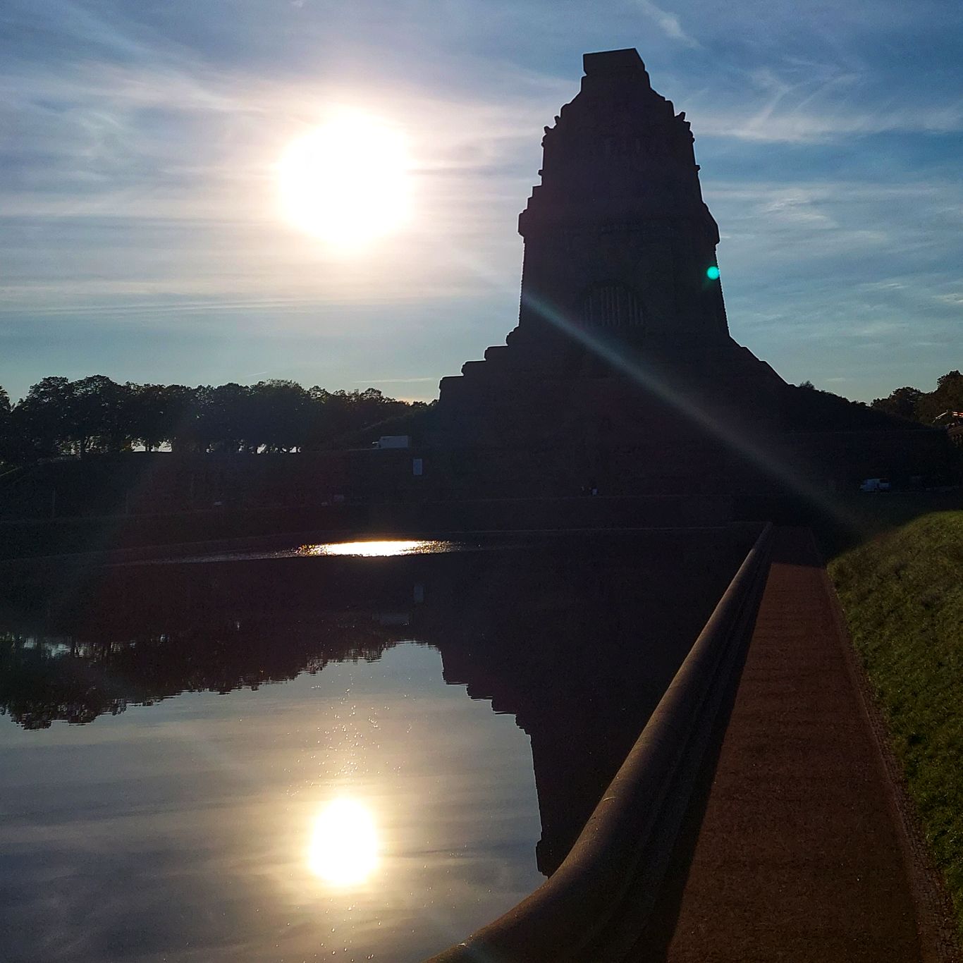 Die aufgehende Sonne neben dem Leipziger Völkerschlachtdenkmal, gespiegelt im davorliegenden Wasserbecken