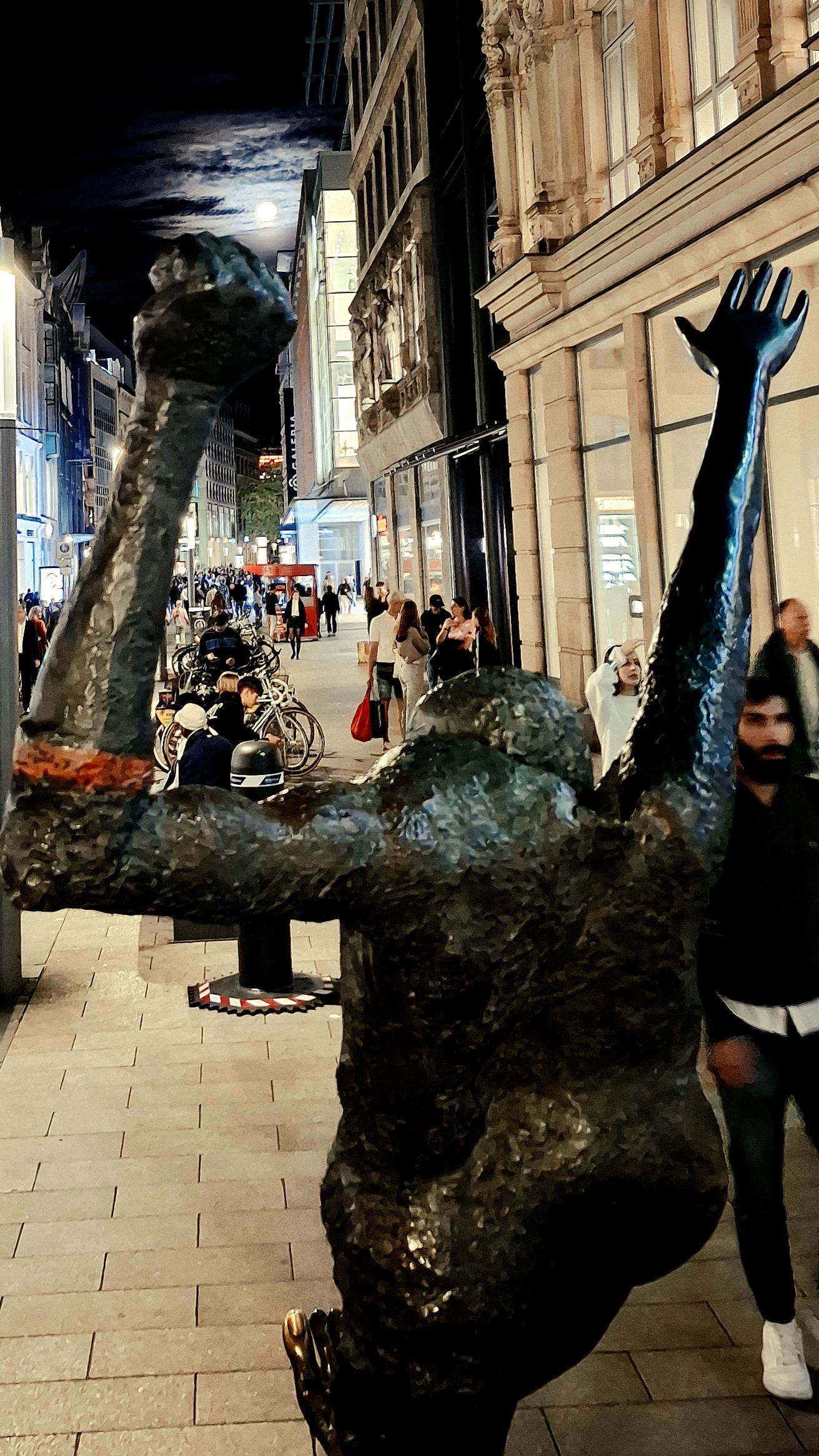 Vonhinten-Foto der Plastik 'Der Jahrhundertschritt' in Leipzig inklusive Fußgängergetümmel zum Ladenschluss an einem  Samstagabend. Über der Faust der Statue schwebt der Vollmond.